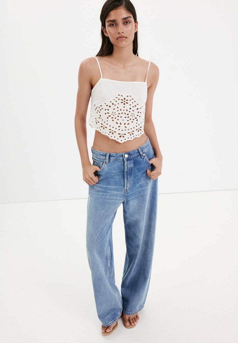Femme debout, mains dans les poches, portant un crop top blanc brodé avec des bretelles fines et un jean bleu ample avec des sandales.
