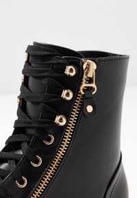 Svart ankelstövel i konstläder, med guldfärgad dragkedja och detaljer, runda guldfärgade öljetter och svarta skosnören.