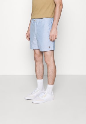 Homme portant un short bleu clair, un t-shirt beige, des chaussettes blanches et des baskets blanches, debout sur un fond uni.