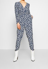 Blommig syntetsdräkt i marinblått med långa ärmar, V-ringning och elastisk midja. Har vita och rosa blomstertryck. Kombination med vita stövletter.