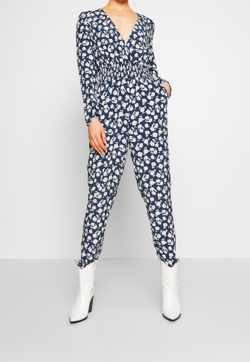 Blommig syntetsdräkt i marinblått med långa ärmar, V-ringning och elastisk midja. Har vita och rosa blomstertryck. Kombination med vita stövletter.