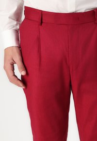 Pantaloni pliați roșii cu o textură netedă, croi ajustat și o talie. O mână stă ușor pe lateralul pantalonilor.