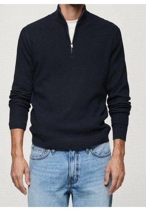 Homme portant un pull côtelé bleu marine à demi-fermeture éclair sur une chemise blanche avec un jean bleu clair, debout, les bras détendus le long du corps.