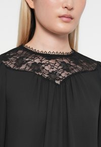 Svart bluse med gjennomsiktig blonde på skuldrene med blomsterdesign og bølget kant, supplert med en flytende design og myk tekstur.