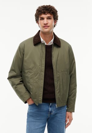 Giovane uomo con capelli ricci che indossa una giacca verde oliva sopra un maglione scuro e jeans azzurri chiari, in piedi con una mano in tasca.