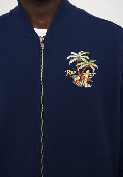Jūras spēku zip-up sporta krekls ar ribotu apkakli. Iezīmēts ar izšuvumu, kurā attēloti palmu koki, sērfa dēlis un vārds "Polo" dzeltenā krāsā.