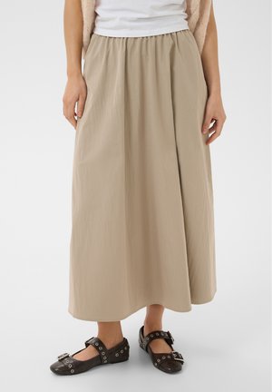 Femme portant une jupe midi beige à taille élastique, un haut blanc et des chaussures noires style Mary Jane avec boucles et œillets.