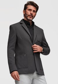 Homme en blazer gris foncé et chemise noire ajustant sa veste, debout devant un fond clair uni.