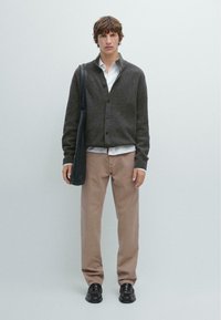 Mørkebrun ribbet cardigan med knapper, hvid skjorte under, lys beige bukser og sorte lædersko. Sort taske hænger fra venstre skulder.