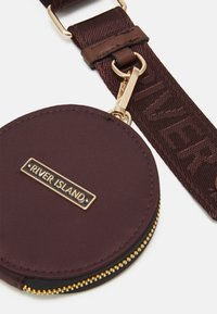 Kulatá burgundská krosová taška z nylonu se zlatým zipem, se značkovou πλαque a strukturovaným popruhem s názvem "RIVER ISLAND."