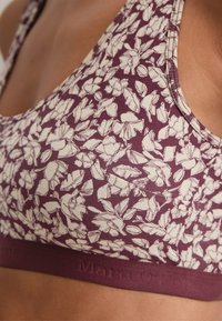 Sport-BH mit floralem Muster in tiefem Burgunderrot und cremefarbenem Blumenprint. Verfügt über ein breites Band mit geprägtem Branding und einem weichen, strukturierten Stoff.