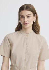 Jonge vrouw met lichtbruin haar, zilveren oorringen en een beige overhemd met korte mouwen, kijkend naar voren.