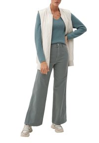 Chaleco sin mangas acolchado de color claro sobre un suéter turquesa, combinado con pantalones sueltos de gris claro y zapatillas beig con detalles verdes.