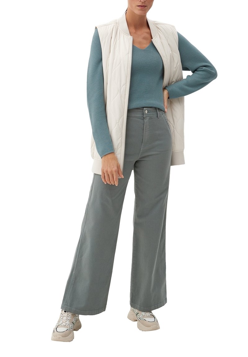 Chaleco sin mangas acolchado de color claro sobre un suéter turquesa, combinado con pantalones sueltos de gris claro y zapatillas beig con detalles verdes.