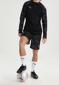 Svart långärmad tränings topp med halvmaterial, kombinerad med svarta mönstrade shorts. Spelaren står på en silvrig fotboll och bär svarta fotbollsskor och vita strumpor.