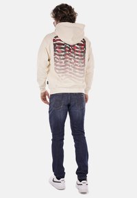 Felpa beige con una stampa grafica di motivi rossi e grigi sul retro. Abbinata a jeans blu e sneakers bianche.