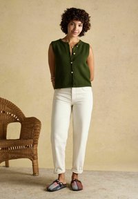 Woman with curly hair stands near wicker chair wearing green sleeveless buttoned vest, white pants, and multicolored slip-on shoes on beige floor.
