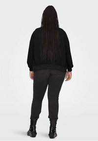 Giacca bomber nera con vestibilità rilassata, polsini e orlo a coste, abbinata a pantaloni scuri e stivaletti neri alla caviglia, vista da dietro.