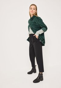 Camicia in corduroy verde sovrapposta a un top a maniche lunghe bianco e verde a righe, abbinata a pantaloni neri a gamba stretta e stivali neri alla caviglia.