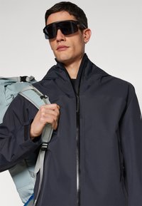 Wasserdichte schwarze Jacke mit hohem Kragen, Reißverschluss und verstellbaren Einstellungen. Begleitet von einem blau-grauen Rucksack. Minimalistisches Design.
