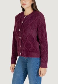 Cardigan finto pelliccia color bordeaux con un motivo a diamante testurizzato, colletto rotondo e cinque bottoni decorativi sulla parte anteriore. Vestibilità sartoriale.