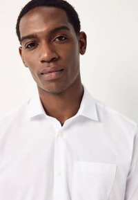 Chemise blanche à boutons avec col pointu, dotée d'une poche unique sur le côté gauche. Tissu lisse et coupe ajustée.