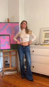 Lace long-sleeve crop top with a high neck; paired with flared blue denim jeans. Wooden easel and colorful artwork in the background.