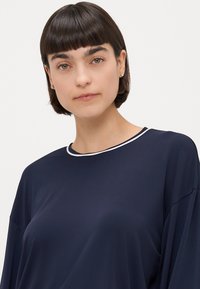 Navy plavi oversize top od mekanog materijala, s okruglim izrezom s bijelim obrubom i širokim rukavima. Glatka tekstura bez uzoraka.