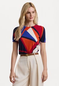 Knit T-shirt featuring a geometric color-block pattern in red, blue, and cream. Short sleeves, round neckline, paired with beige trousers.