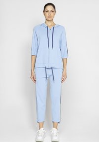 Jasnoniebieski dres wykonany z miękkiej bawełny. Posiada zapinaną na zamek kurtkę z kapturem, dwie przednie kieszenie i sznurki; dopasowane spodnie z bocznymi paskami.