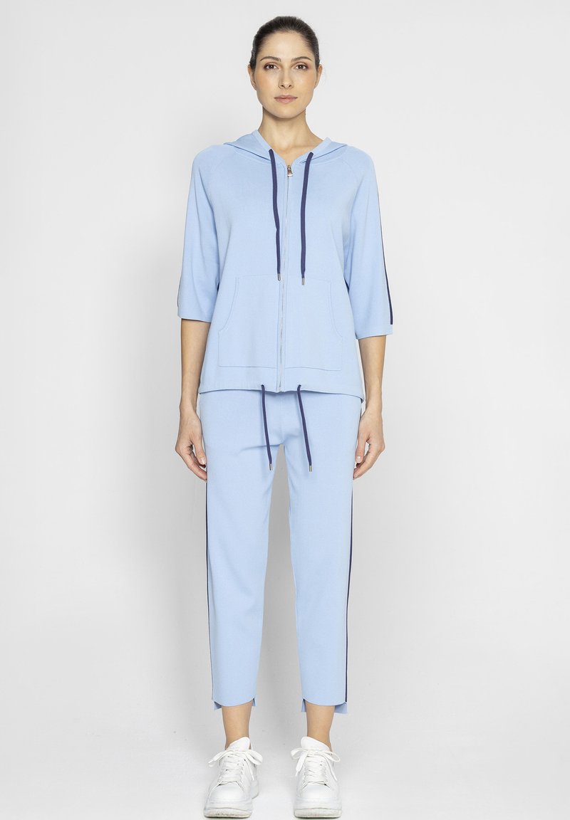 Jasnoniebieski dres wykonany z miękkiej bawełny. Posiada zapinaną na zamek kurtkę z kapturem, dwie przednie kieszenie i sznurki; dopasowane spodnie z bocznymi paskami.