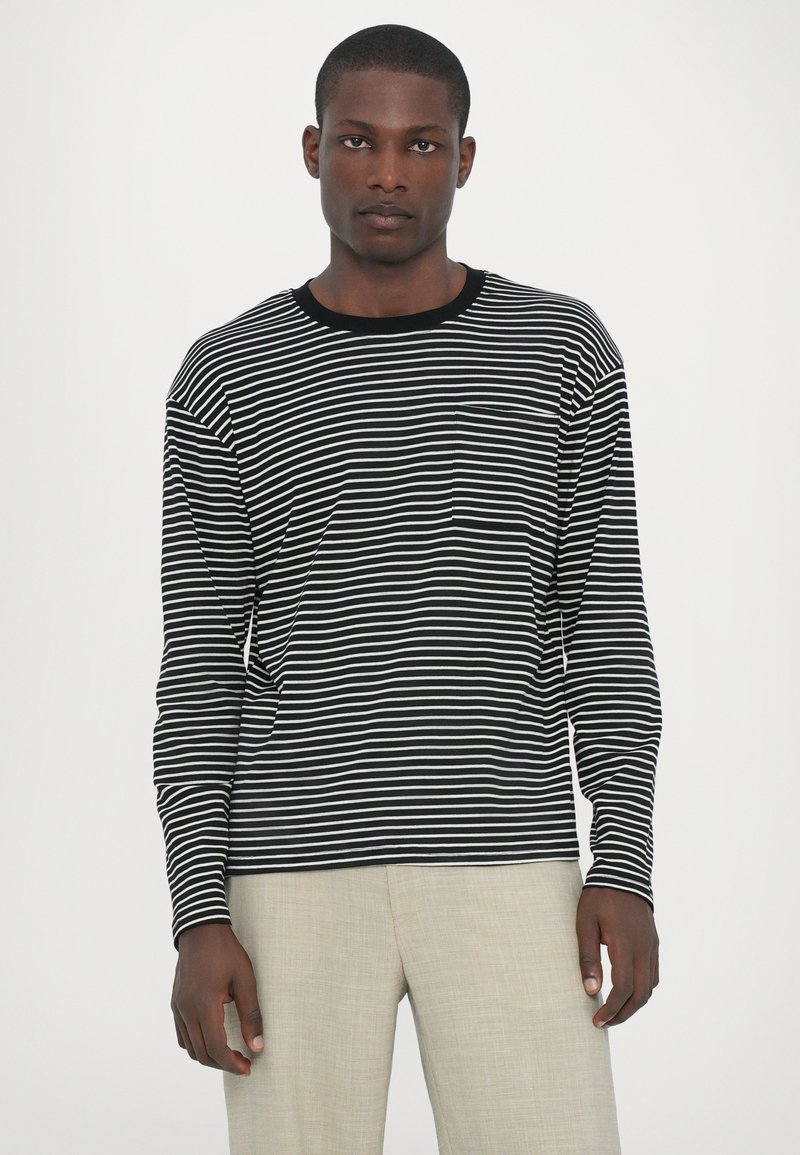 Schwarz-weiß gestreifes Langarmshirt mit Rundhalsausschnitt und einer Brusttasche, getragen mit hellbeigen Hosen. Glatte Stoffstruktur.