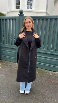 Fekete, dupla soros kabát széles hajtókával, fekete, testhezálló felső fölött viselve, világoskék, bővülő szárú farmerral és fehér tornacipővel párosítva.