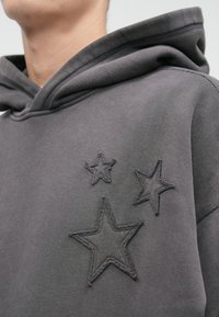 Casaco de capuz cinza com três designs de estrelas bordados em tamanhos variados, costura texturizada e uma silhueta clássica com capuz ajustável.
