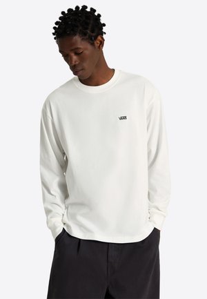 Mann mit Händen in den Taschen, der ein weißes langärmeliges Vans-Shirt und schwarze Hose trägt, blickt nach unten, vor schlichtem hellem Hintergrund.