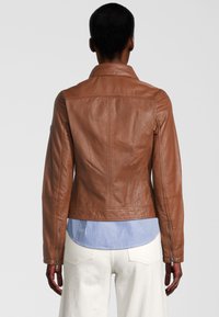 Veste en cuir marron au design ajusté, avec des coutures visibles et un petit col. Les manchons zippés et l'ourlet d'une chemise rayée sont partiellement visibles en dessous.