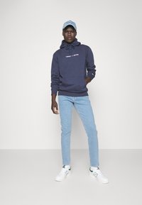 Sudadera azul marino con un bolsillo frontal y el logo blanco "TOMMY JEANS", combinada con unos pantalones vaqueros de un azul claro y zapatillas blancas con detalles verdes.