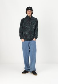 Fleece genser i mørkegrønn og svart virvlende mønster, halv-zip krage, kombinert med lyseblå baggy jeans og svarte joggesko. Svart caps.