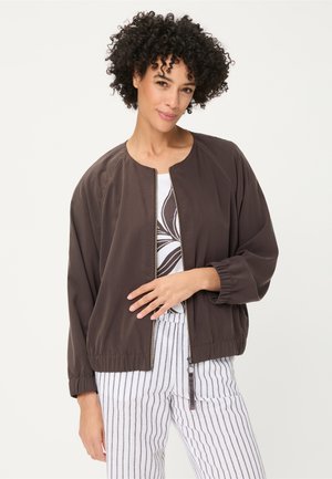 Woman with curly hair wearing a dark brown zip-up jacket, white shirt with a black abstract design, and white striped pants, standing against white background.
