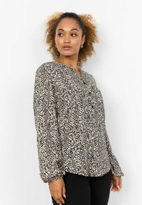 Långärmad blus i beige med ett svart abstrakt mönster, med V-halsringning och elastiska ärmslut. Slät, lättviktsmaterial.