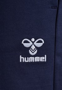 Navyblauwe stof met een witte geborduurde logo met een bijontwerp en de merknaam "hummel" eronder, tegen een gestructureerde achtergrond.