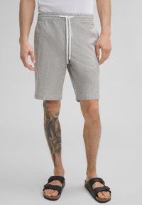 Gestreifte graue und weiße Shorts mit einem elastischen Bund und einem weißen Kordelzug, ausgestattet mit seitlichen Taschen und einem lockeren Schnitt. Getragen mit schwarzen Sandalen.