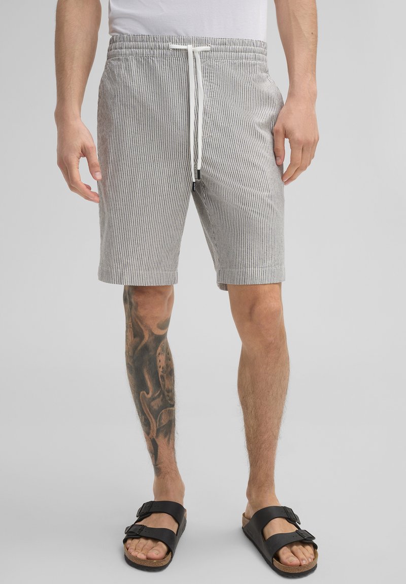 Gestreifte graue und weiße Shorts mit einem elastischen Bund und einem weißen Kordelzug, ausgestattet mit seitlichen Taschen und einem lockeren Schnitt. Getragen mit schwarzen Sandalen.