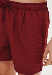 Burgundowe szorty kąpielowe z elastycznym pasem, zaciąganym sznurkiem i bocznymi kieszeniami. Lekki materiał o gładkiej teksturze.