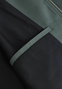 Gros plan d’un tissu noir et vert foncé avec un motif hexagonal et une bordure verte près d’une fermeture éclair argentée sur un vêtement.