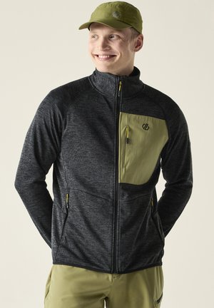 Junger Mann lächelt, trägt eine dunkelgraue Jacke mit Reißverschluss und olivgrüner Brusttasche sowie eine olivgrüne Mütze, steht vor hellem Hintergrund.