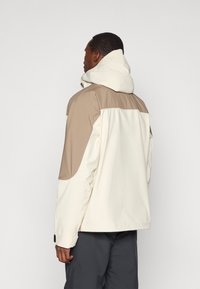 Casaco com capuz em bege e creme, com design de aba, tecido texturizado e punhos ajustáveis, exibido pela parte de trás contra um fundo simples.