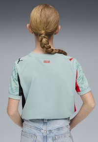 Maglia sportiva azzurro chiaro con maniche corte, caratterizzata da dettagli rossi, lati neri e una manica a fantasia. "MILAN" in rosso sulla parte posteriore del collo.