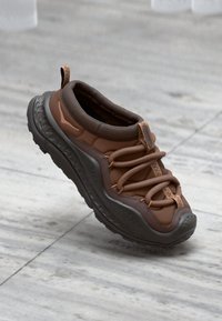 Zapatilla marrón con parte superior acolchada, suela gruesa gris y correas texturizadas. El diseño incluye una mezcla de materiales suaves y gomosos.