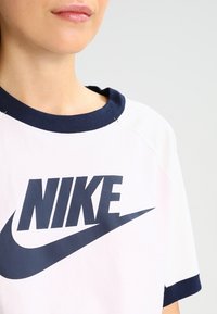 Person som bär en vit Nike t-shirt med marinblå krage och ärmslut, där Nike-logotypen och swoosh syns på bröstet.